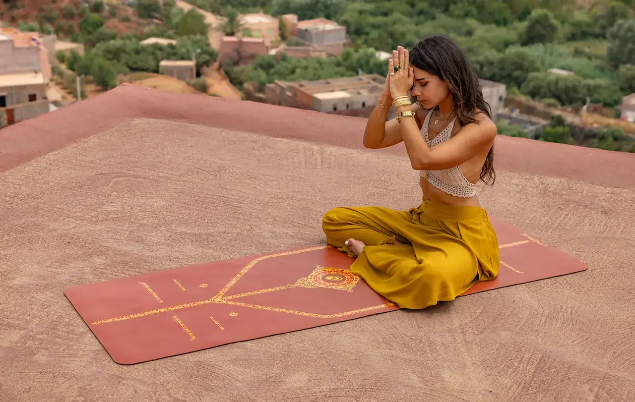 Mujer en postura de loto en una esterilla de yoga, con las manos juntas en gesto de namasté. El paisaje de fondo es un pueblo con casas de adobe y vegetación.