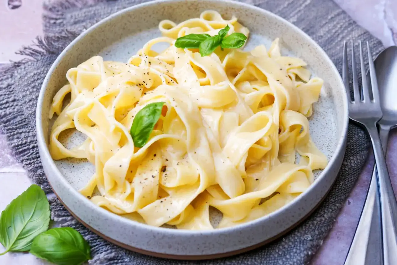 Cremiges Pasta Alfredo mit frischem Basilikum und schwarzem Pfeffer, serviert in einer grauen Sch&uuml;ssel auf einem rustikalen Tuch.