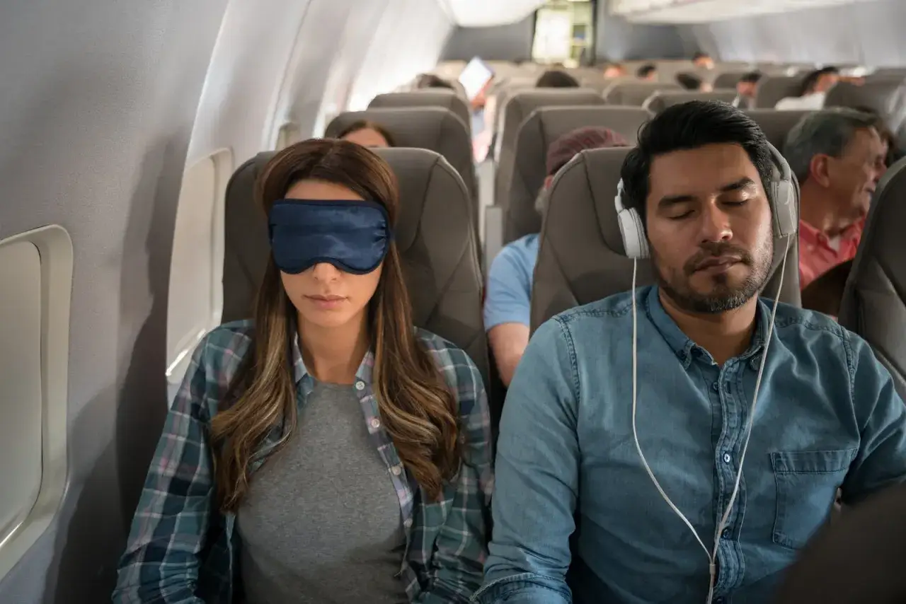 Två passagerare försöker sova på ett flygplan, en med sovmask och den andra med hörlurar. De kämpar mot jetlag usa till sverige.