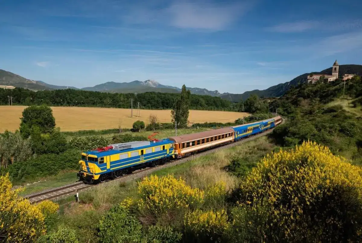 Un tren azul y amarillo recorre un paisaje verde con campos dorados y un pueblo en la colina.