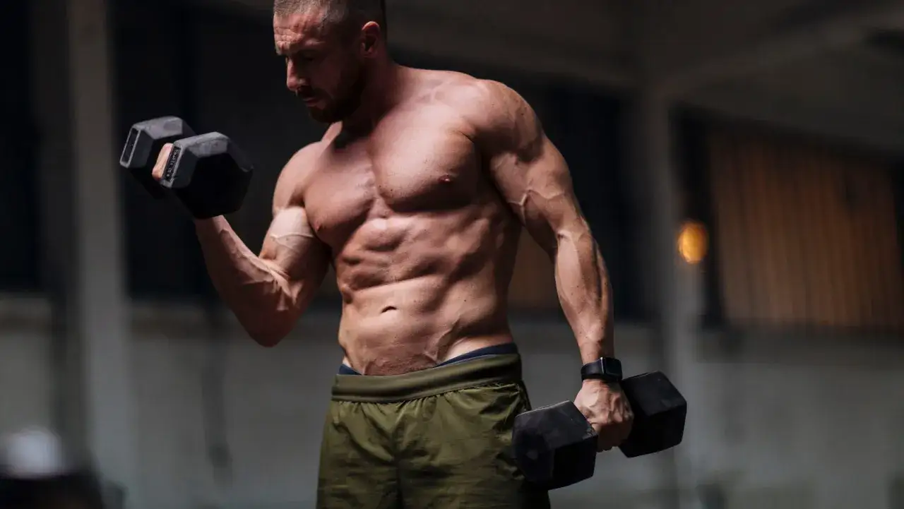 Mężczyzna z wyrzeźbionym torsem wykonuje ćwiczenia na biceps z hantlami.