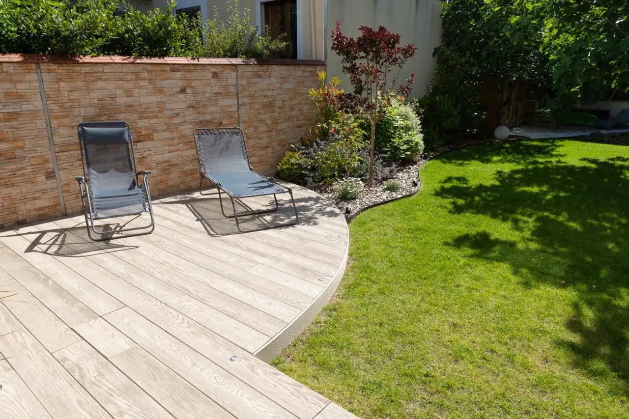 Am&eacute;nagement ext&eacute;rieur : terrasse en bois, deux chaises longues, mur de pierres et pelouse verdoyante. Un espace d&eacute;tente parfait.
