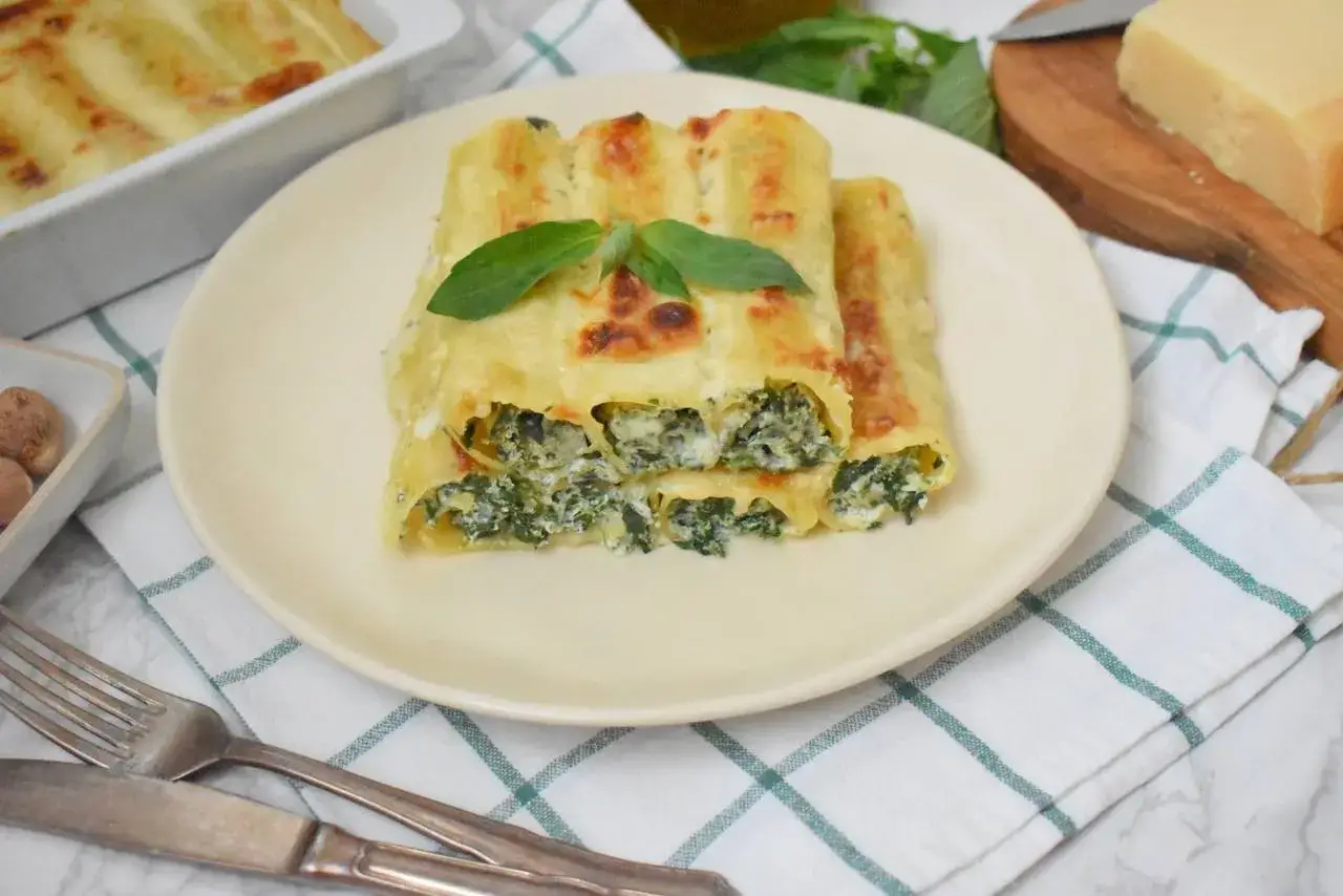 Pyszne cannelloni ze szpinakiem, zapieczone pod beszamelem i serem, podane na talerzu z listkiem bazylii.