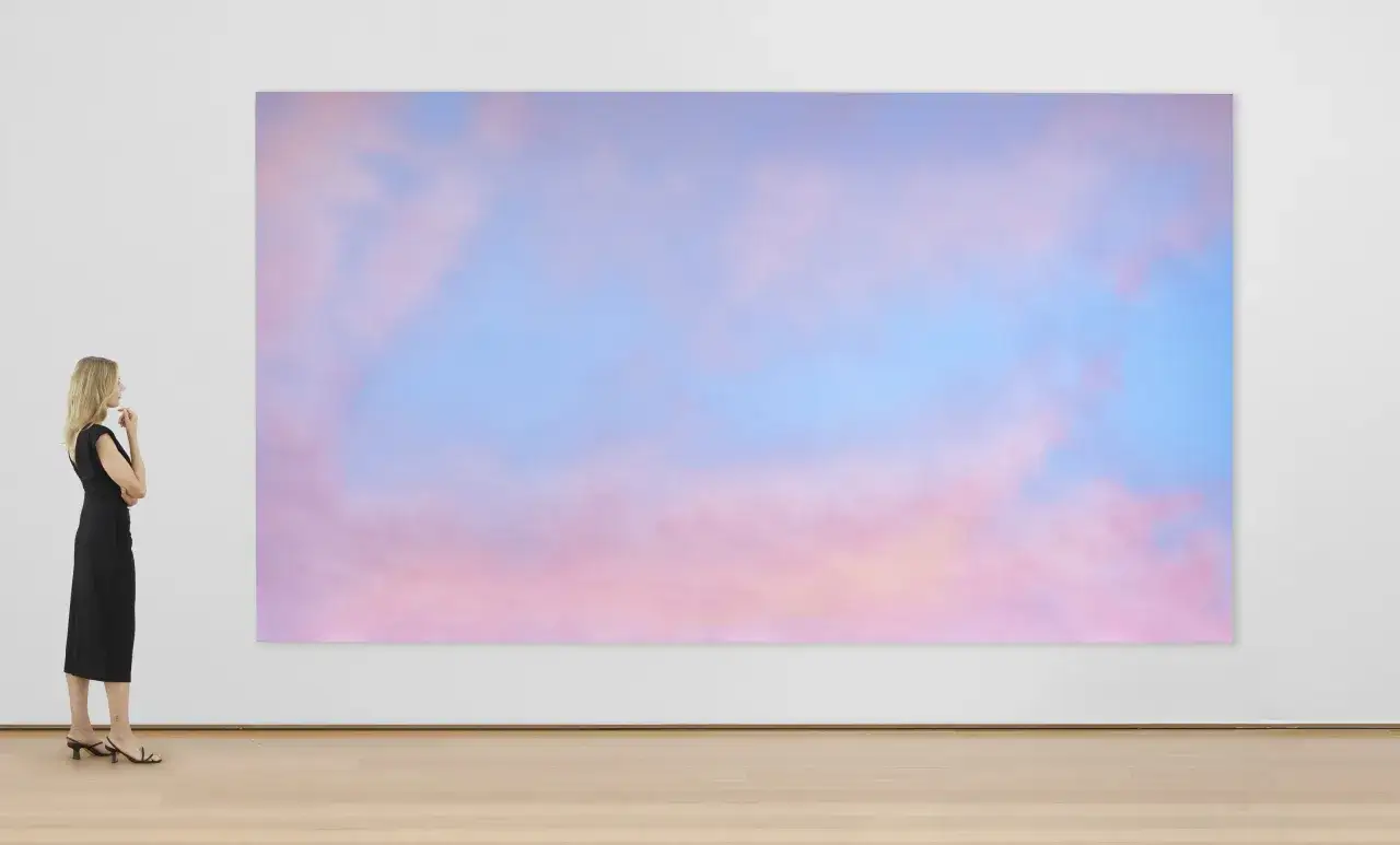 A woman in a black dress contemplates a large, abstract painting of pink and blue clouds, reminiscent of Alex Israel's signature style.
