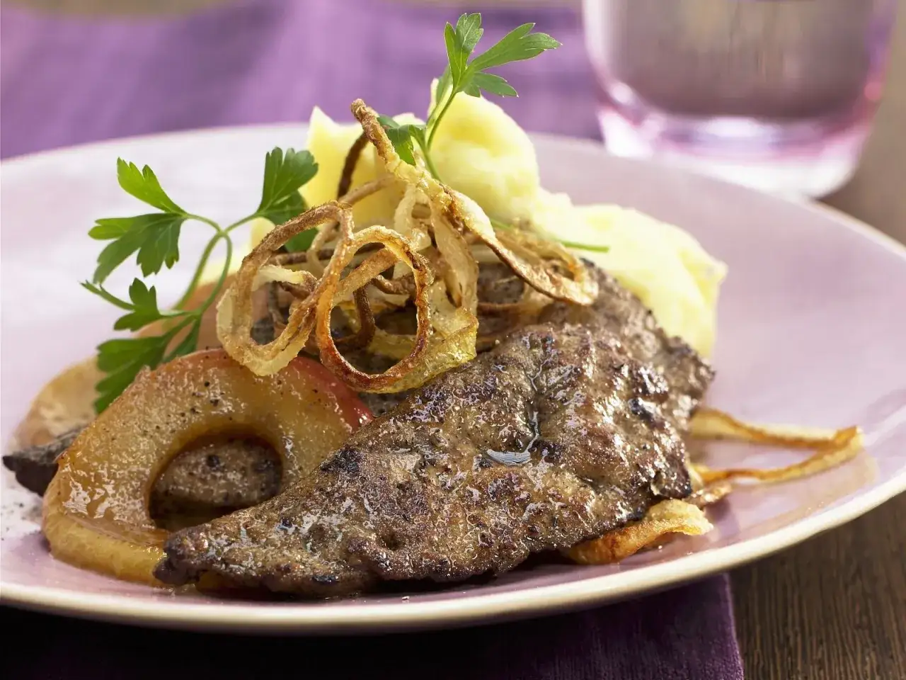 Gebratenes Lammleber-Rezept mit Apfelringen, Kartoffelpüree und knusprigen Zwiebelringen auf einem rosa Teller.