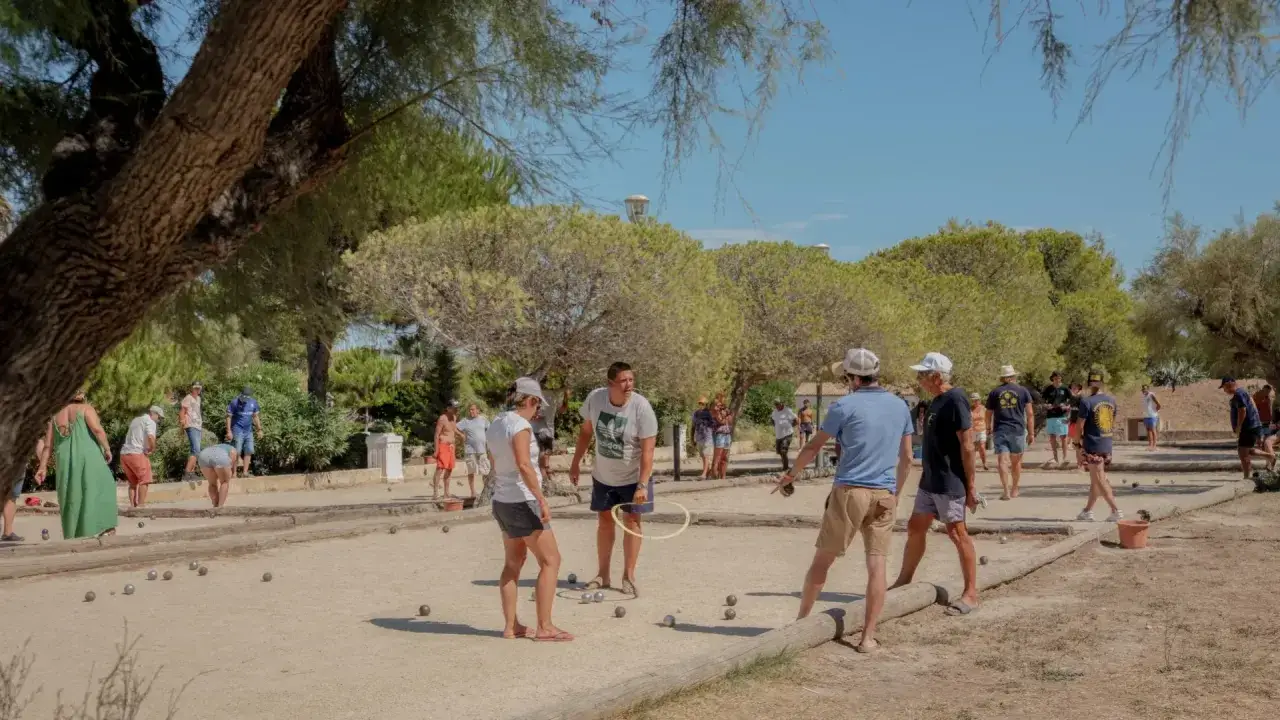 Des joueurs s'affrontent dans une partie de pétanque animée, lançant des boules sur le terrain sablonneux sous un ciel bleu.