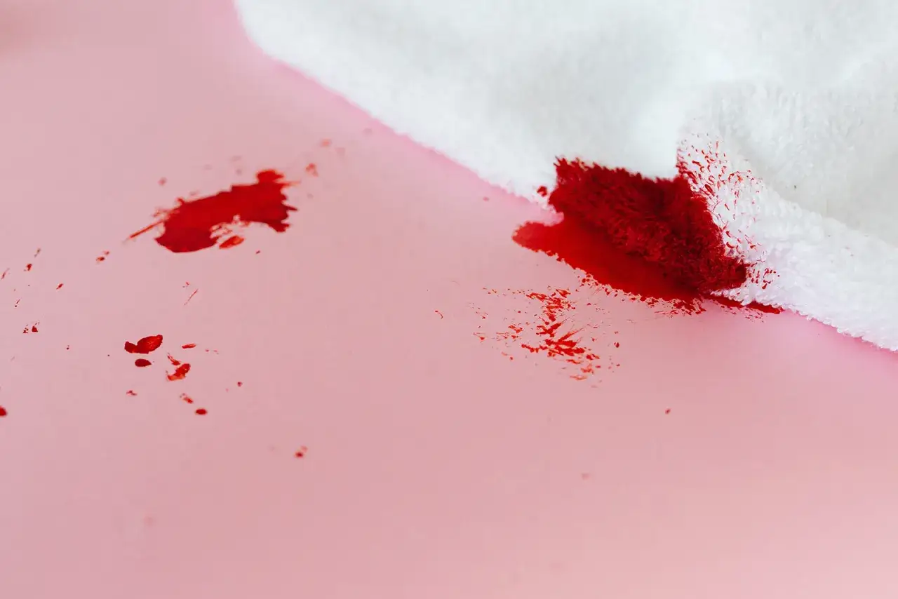 Rode bloedvlekken op roze ondergrond, met een witte doek die de vlekken opneemt. Oma weet raadt voor bloedvlekken verwijderen.