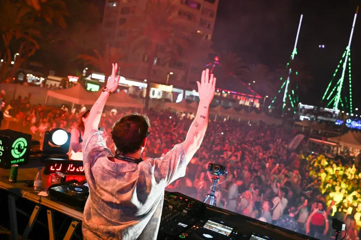 DJ con le braccia alzate su un palco a Ibiza, la folla balla al ritmo della sua canzone.