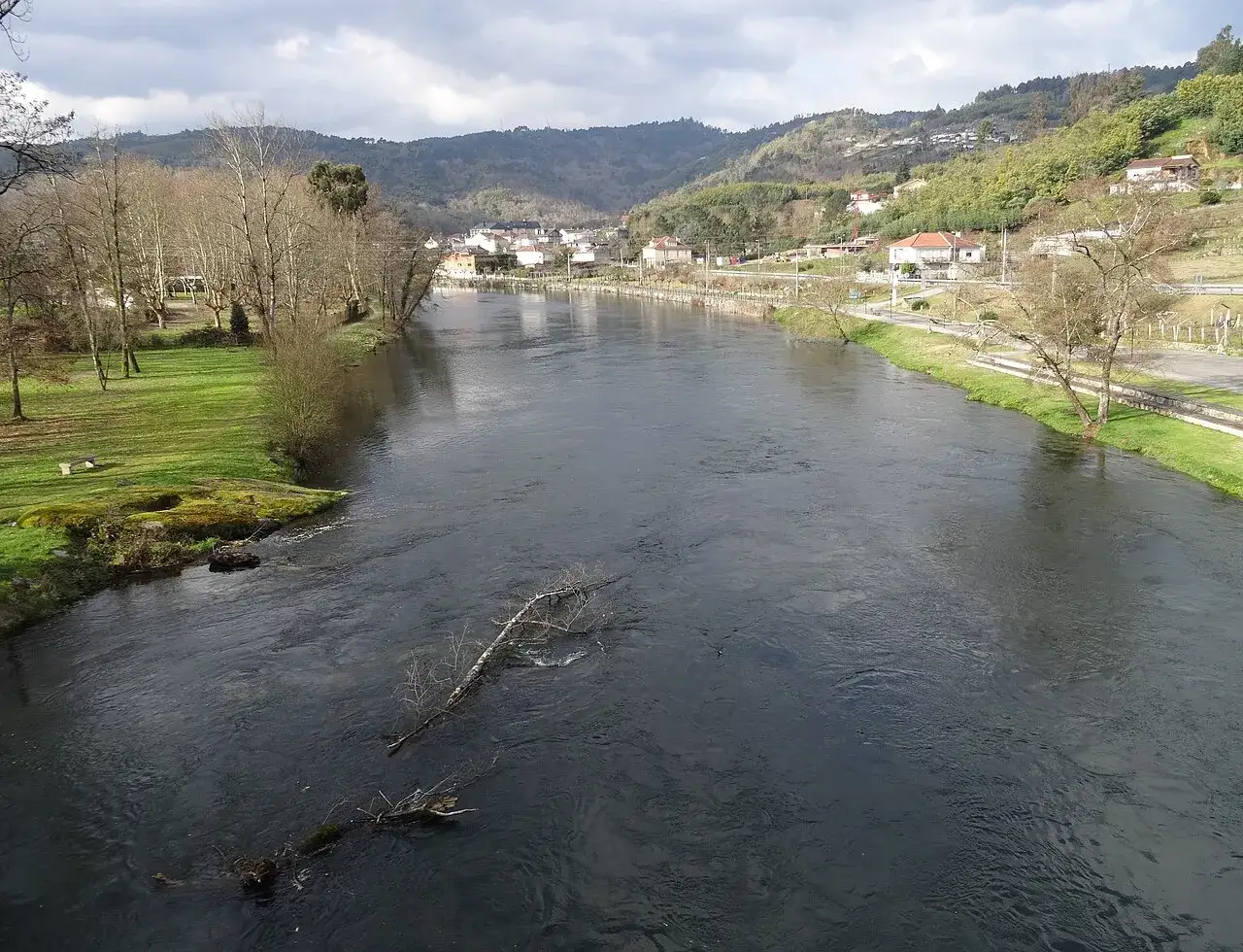 El río fluye sereno cerca de la playa fluvial Leiro, con árboles desnudos y casas en la ladera.