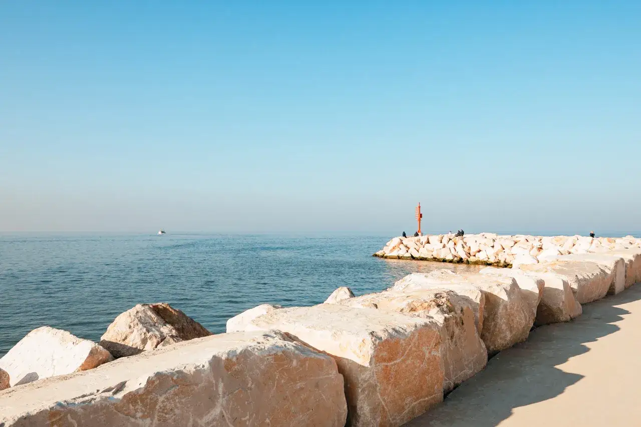 Scogliera di igea marina bellaria con un faro rosso e persone sedute. Mare calmo e cielo azzurro.