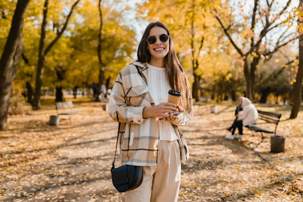 Uśmiechnięta kobieta w beżowym, kratowanym swetrze i spodniach, z kubkiem kawy w dłoni. Idealny casual style damski na jesienny spacer.