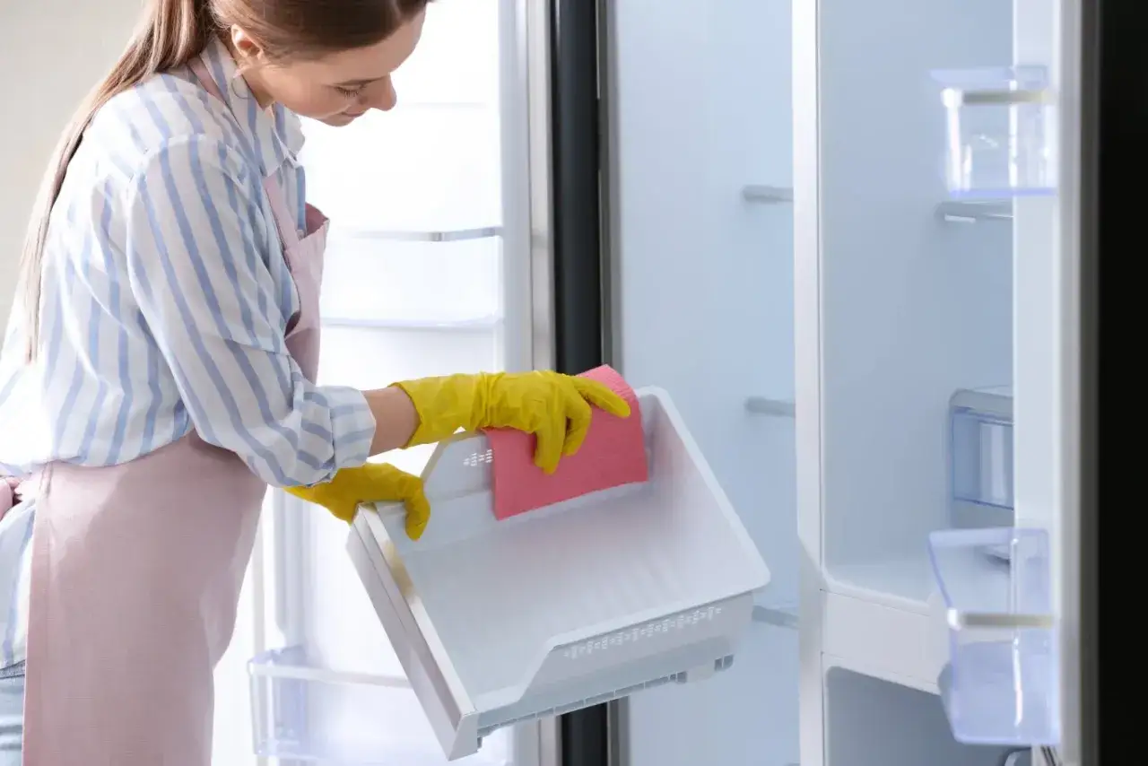 Kobieta w żółtych rękawiczkach czyści szufladę lodówki, szukając odpowiedzi, dlaczego lodówka śmierdzi mimo regularnego mycia i wyrzucania jedzenia.