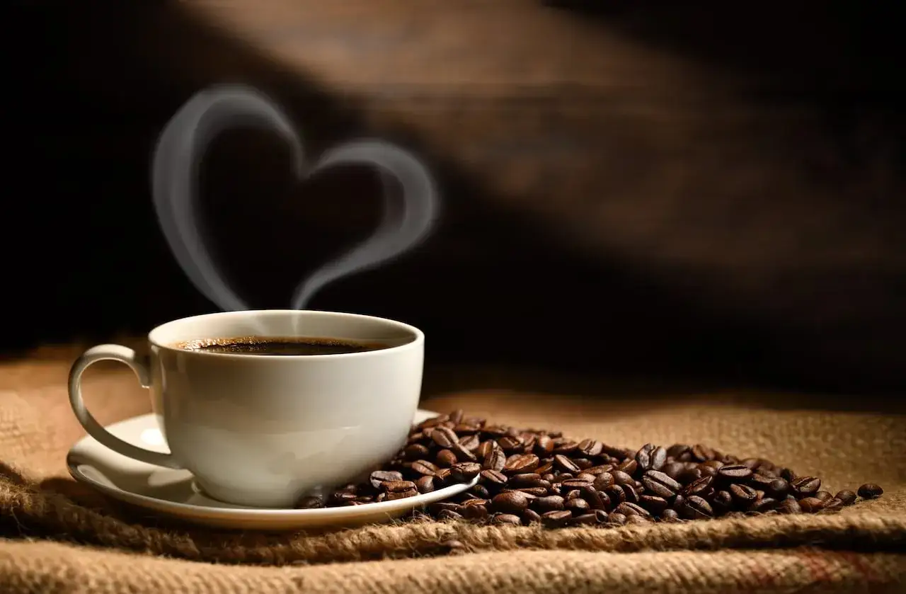 Una taza de café humeante con vapor en forma de corazón, rodeada de granos de café. Dicen que el café es afrodisiaco.