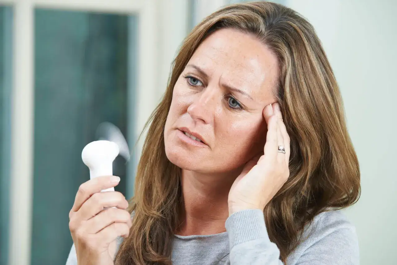 Kobieta z ręką na skroni, trzymająca mały wiatraczek. Objawy menopauzy, depresja, nerwica.