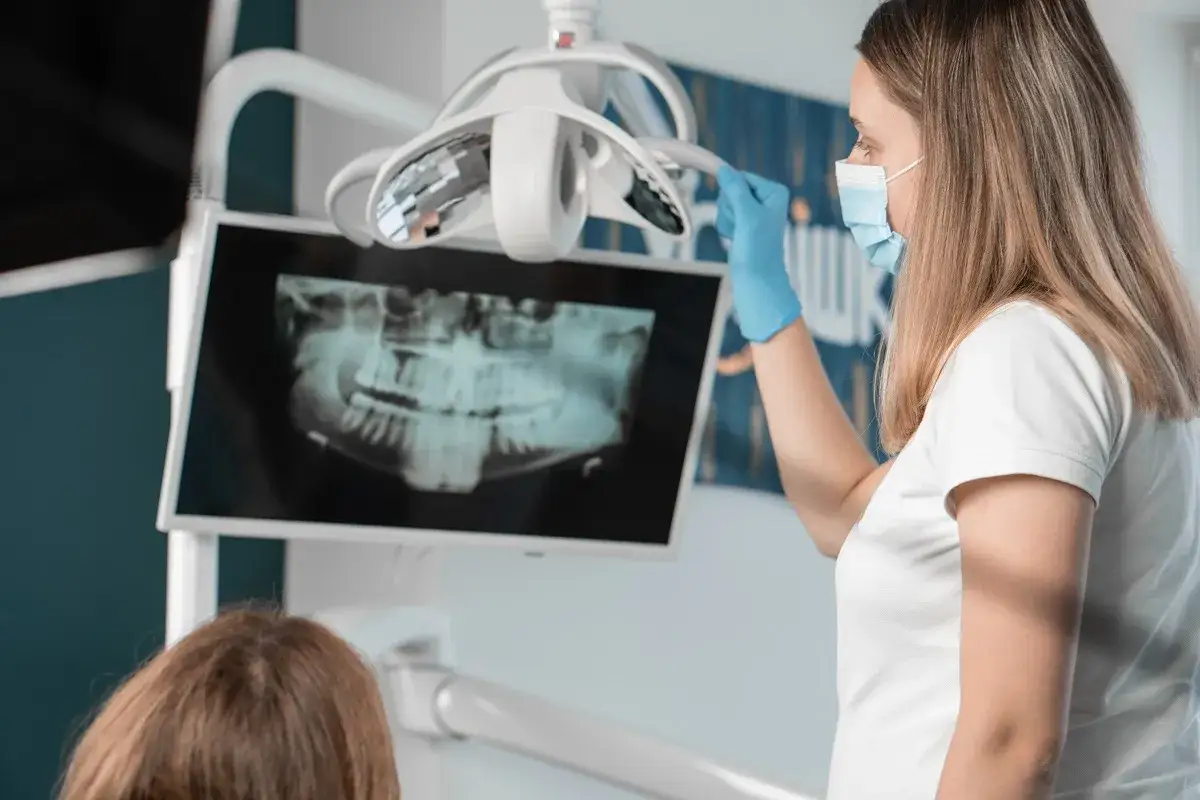 Dentystka w masce i rękawiczkach pokazuje pacjentowi zdjęcie rtg zębów na monitorze.