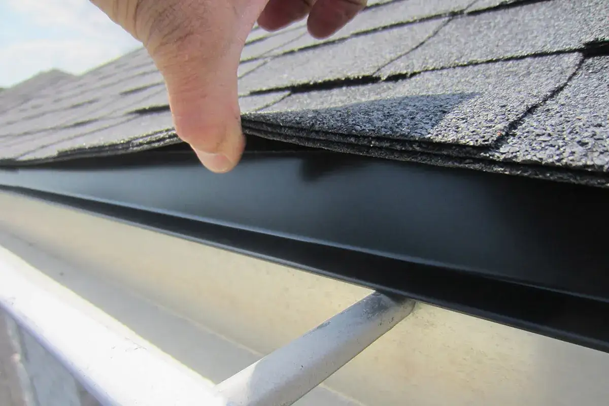 A hand lifts shingles to reveal the black drip edge, protecting the roof and gutter from water damage.