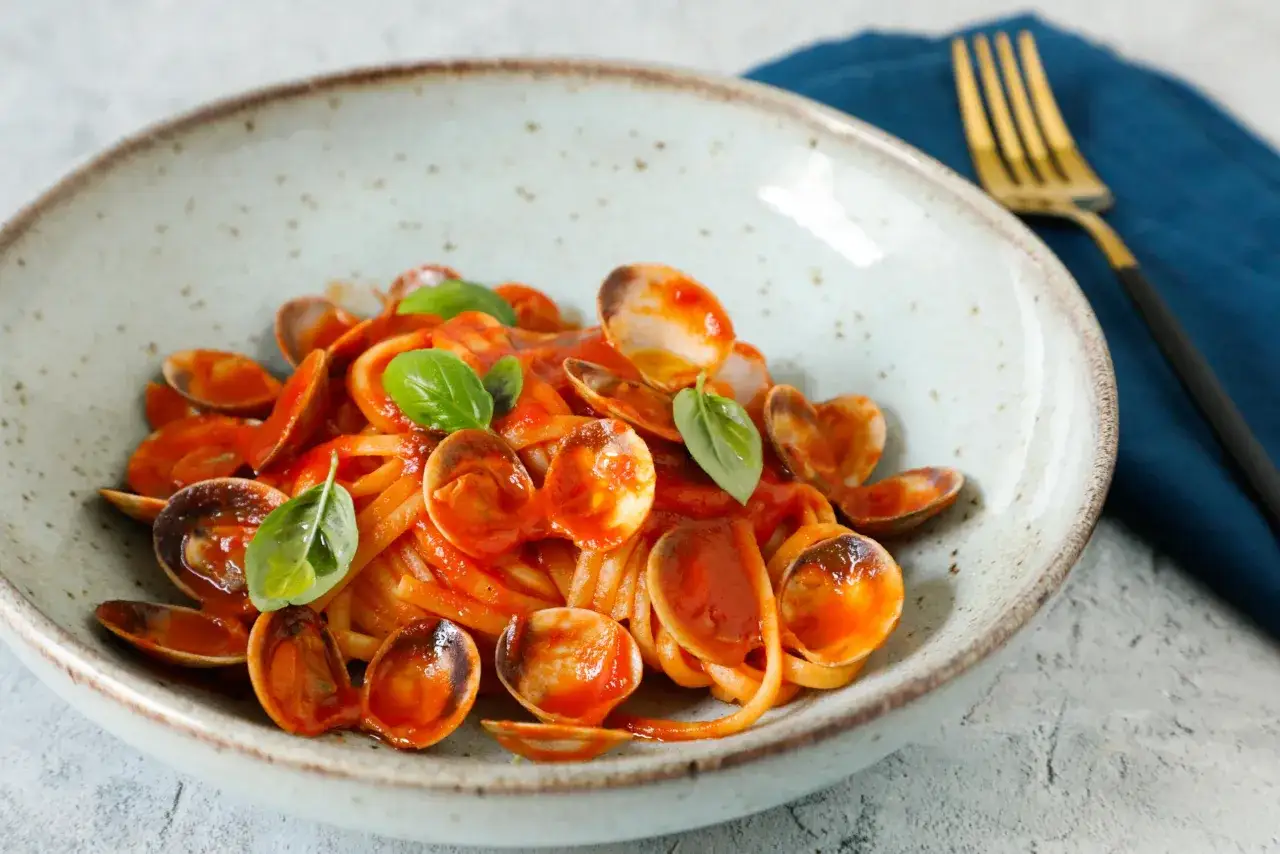 Spaghetti con vongole surgelate, conditi con sugo rosso e basilico fresco, serviti in una ciotola rustica.