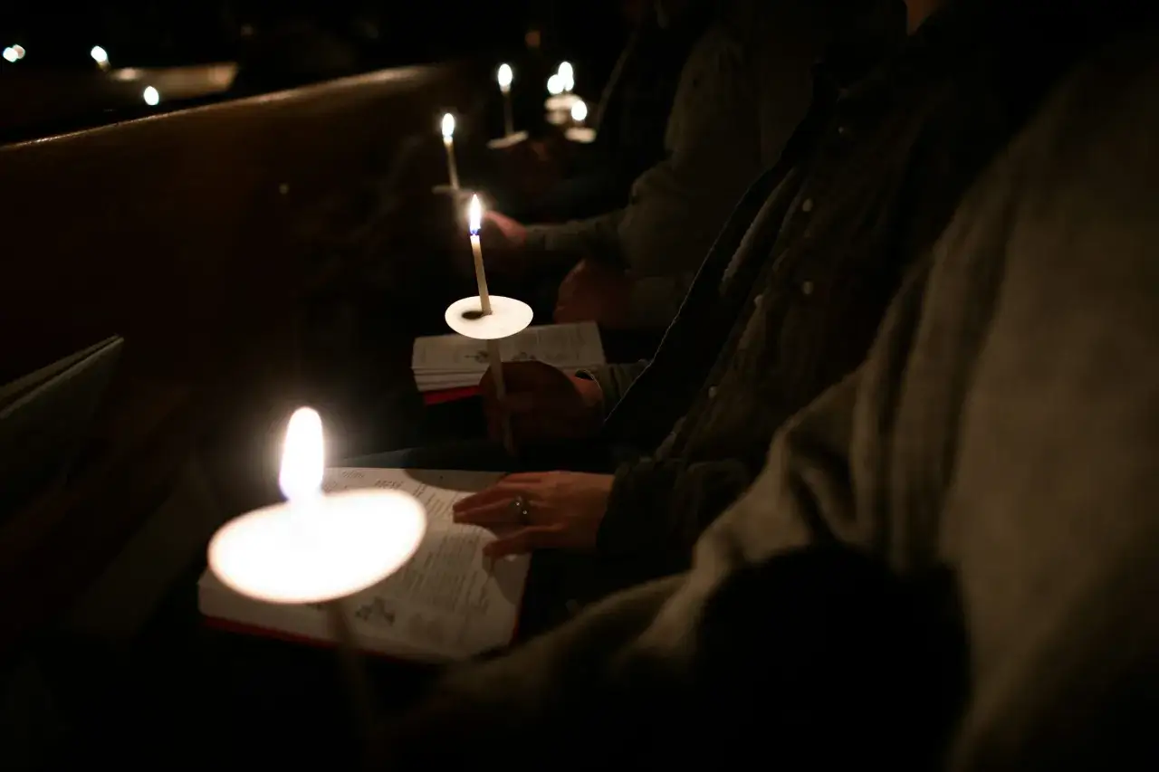 Kerzenlicht erhellt eine **Vigil Kirche**. Menschen halten brennende Kerzen und lesen in B&uuml;chern.