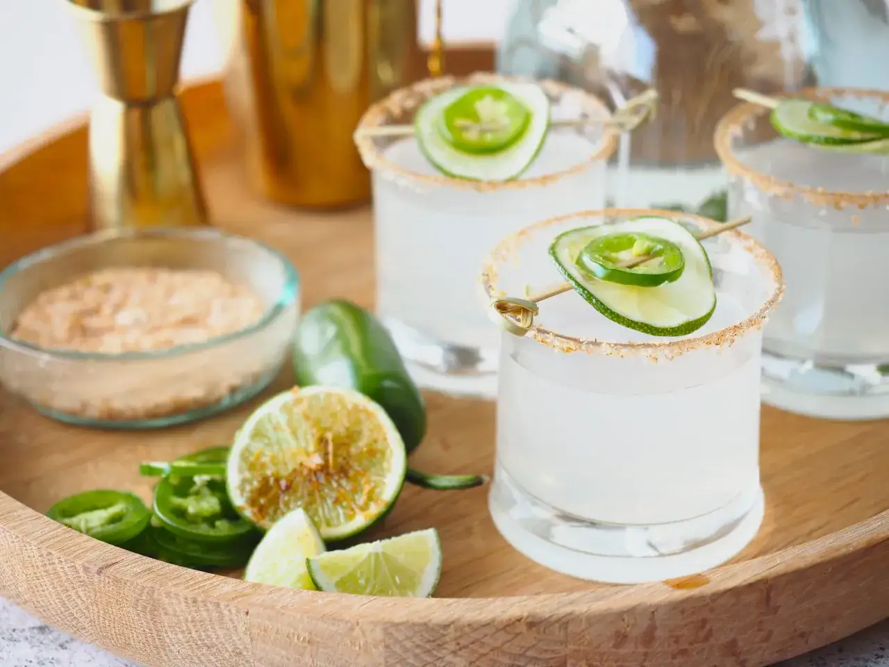Ein scharfes Margarita Rezept: Gläser mit Salzrand, garniert mit Limette und Jalapeño. Zutaten wie Limetten, Jalapeños und Salz sind auf einem Holzteller arrangiert.