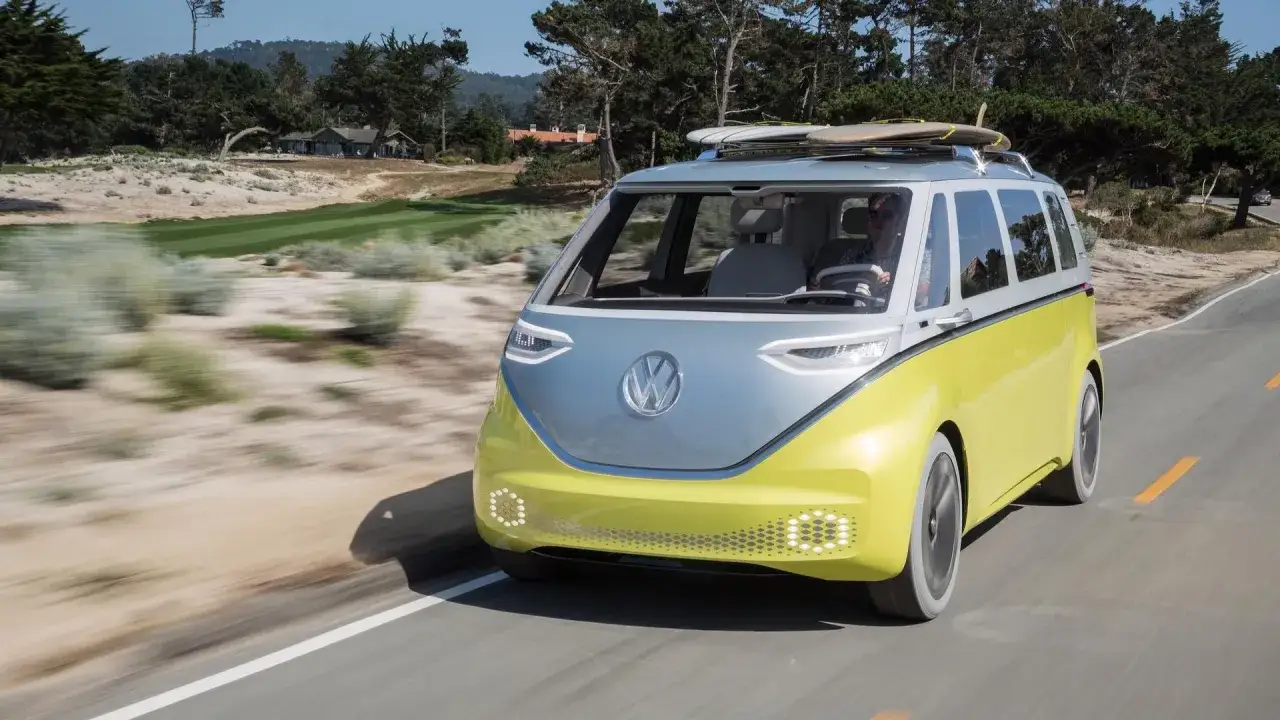 Żółto-szary minivan Volkswagen z deskami surfingowymi na dachu jedzie drogą przy wybrzeżu.