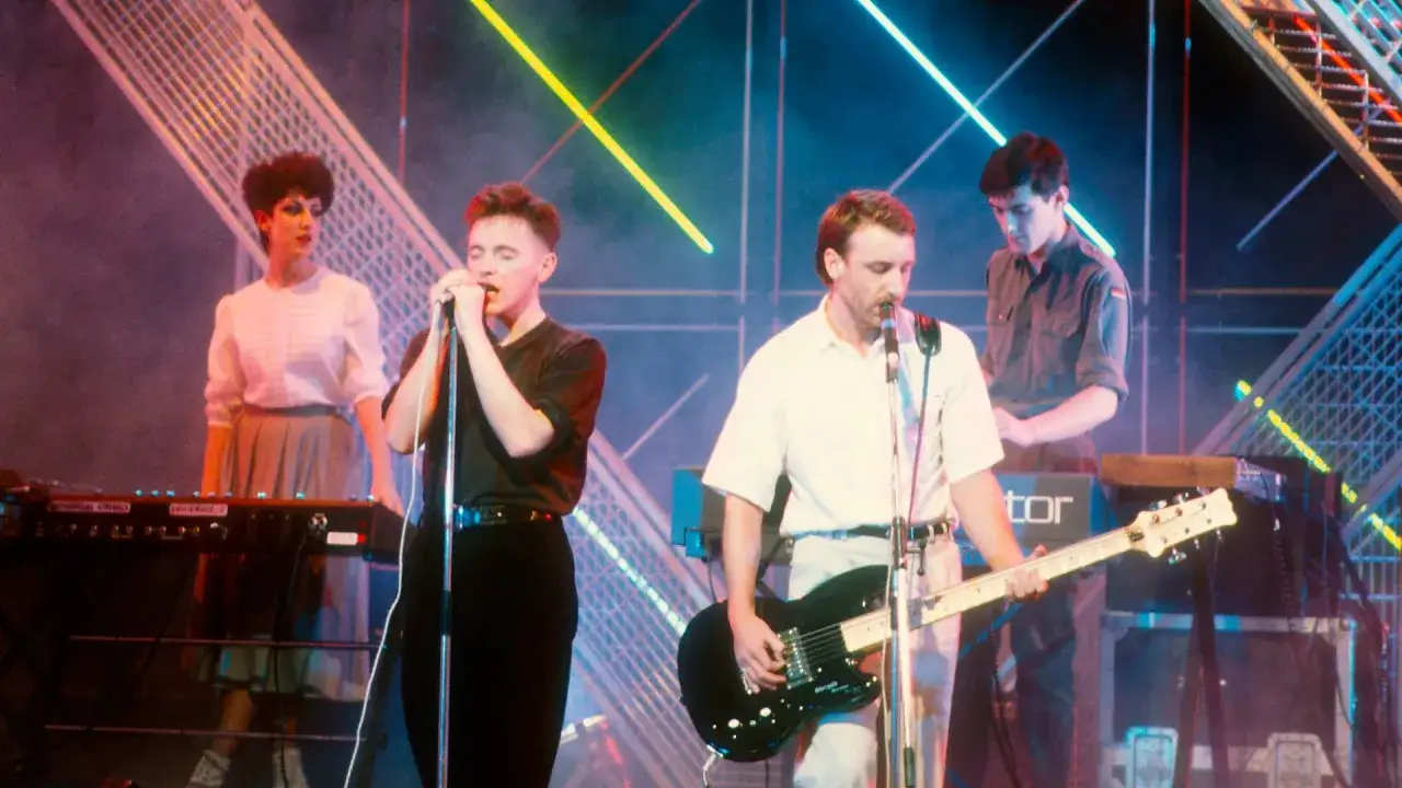 Zespół New Order wykonuje swoje utwory na żywo. Wokalista śpiewa do mikrofonu, gitarzysta gra na gitarze, a klawiszowiec obsługuje syntezator.