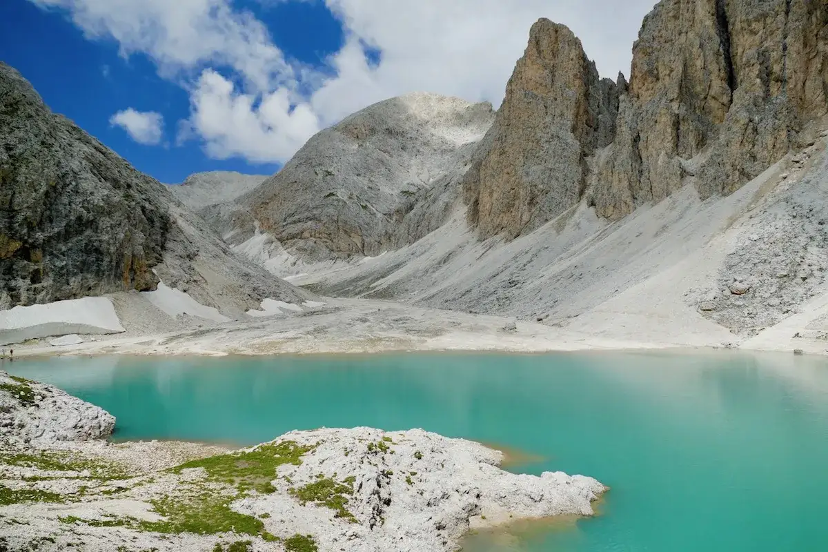 Il **lago di Antermoia** riflette le imponenti vette rocciose sotto un cielo azzurro con nuvole bianche.
