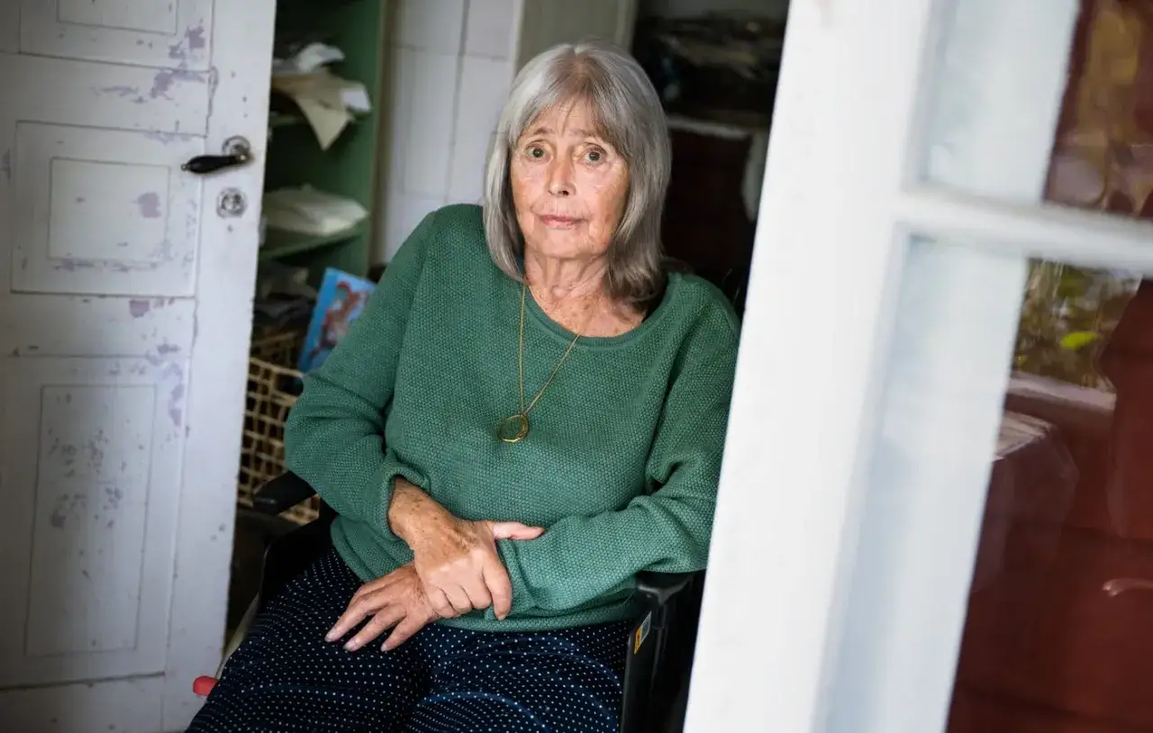 Agneta Pleijel ung, en &auml;ldre kvinna med gr&aring;tt h&aring;r, sitter i en rullstol i ett rum med en &ouml;ppen d&ouml;rr. Hon b&auml;r en gr&ouml;n tr&ouml;ja och ett halsband.