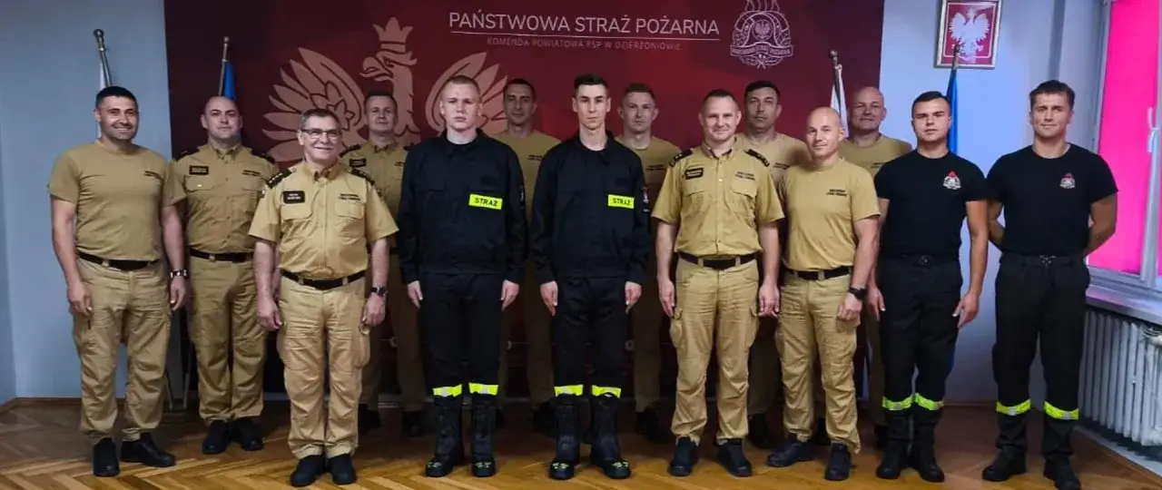 Gotowa do akcji rota strażacka pozuje do zdjęcia. Mundury w kolorze khaki i czerni świadczą o ich gotowości do służby.