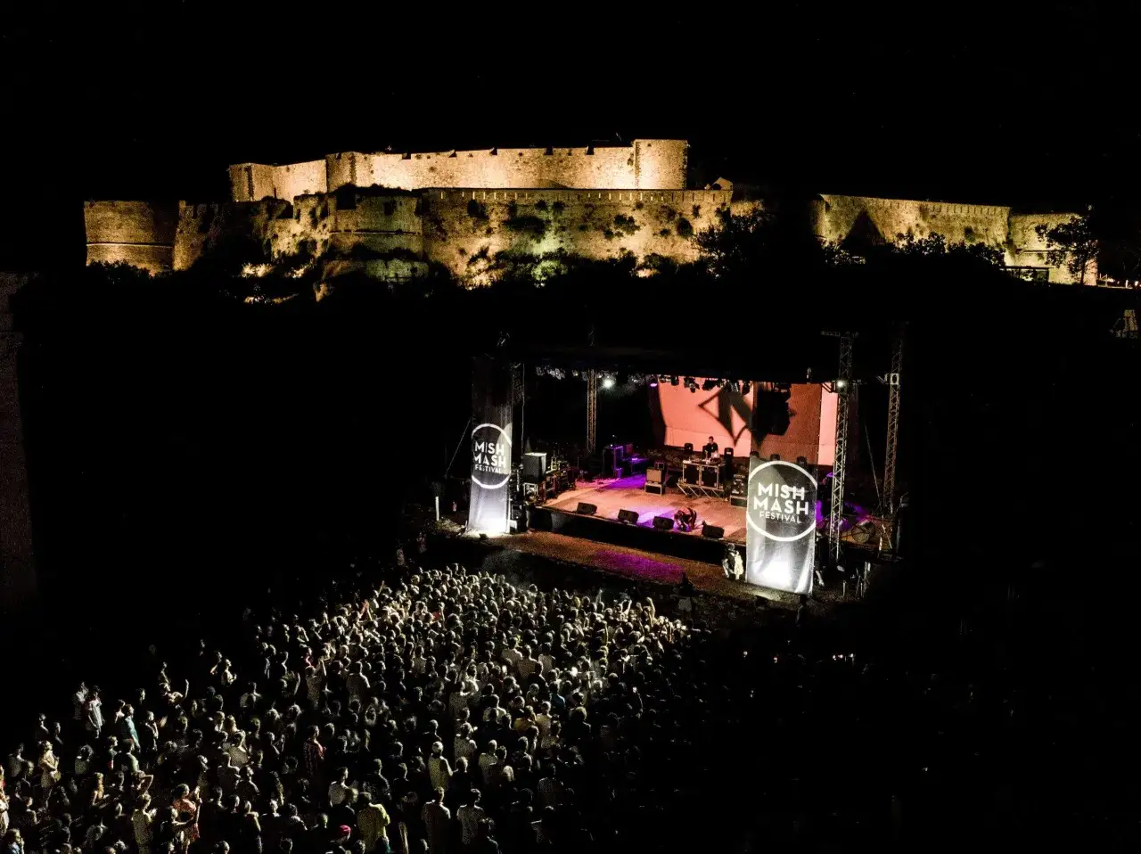 Un festival notturno con un palco illuminato e una folla danzante, sullo sfondo un antico castello. Forse i dj siciliani famosi stanno facendo ballare tutti!