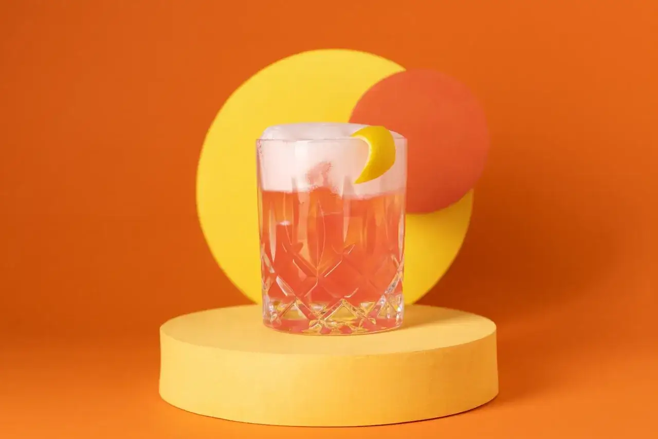 Ein erfrischender Aperol Sour mit schaumiger Haube und Zitronenzeste, pr&auml;sentiert auf einem gelben Podest vor orangefarbenem Hintergrund mit abstrakten Kreisen.