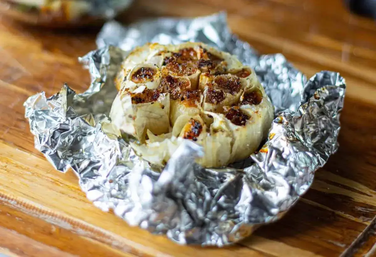 Une t&ecirc;te d'ail confit au four, dor&eacute;e et caram&eacute;lis&eacute;e, envelopp&eacute;e dans du papier d'aluminium froiss&eacute; sur une planche en bois.