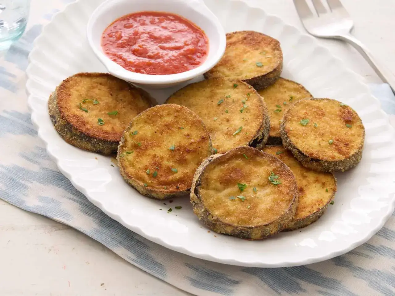 Krispigt fritera aubergine p&aring; en vit tallrik med en sk&aring;l tomats&aring;s. Garnerad med persilja.