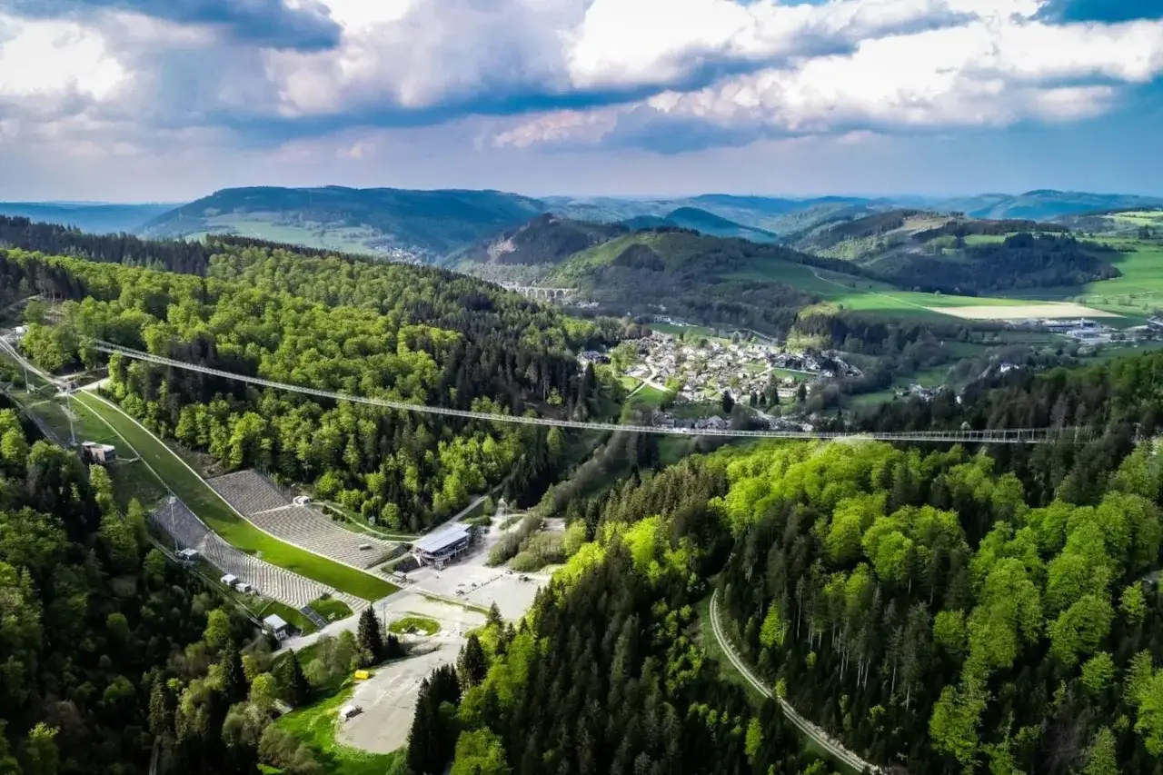 Der Willingen Skywalk &uuml;berspannt ein gr&uuml;nes Tal mit W&auml;ldern und einer kleinen Stadt. Eine Skisprungschanze ist links zu sehen.