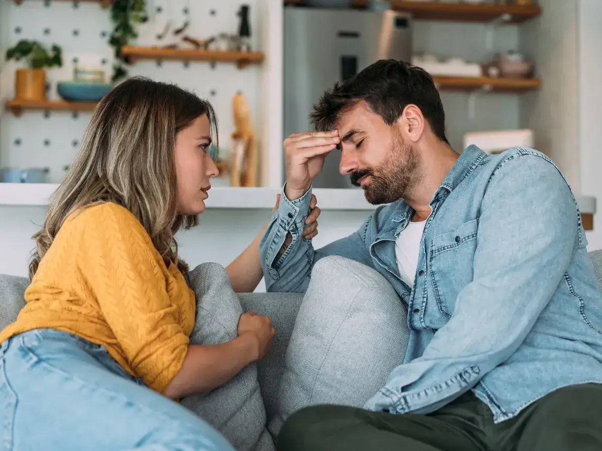 Pareja discutiendo en el sof&aacute;. Ella le habla con seriedad, &eacute;l se lleva la mano a la frente, agobiado por el chantaje emocional en la pareja.