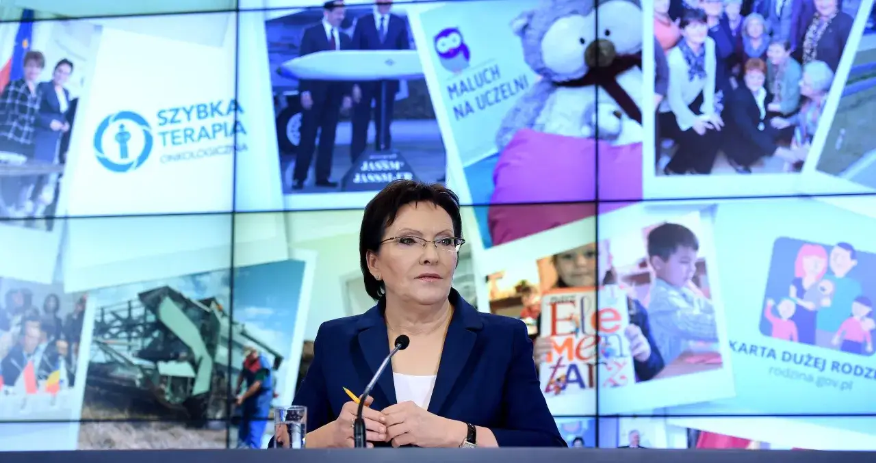 Ewa Kopacz, premier, przemawia na tle ekranów z grafikami o tematyce społecznej i medycznej.
