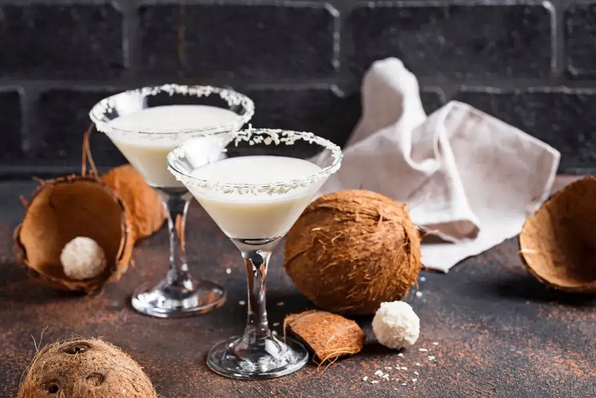 Zwei elegante Cocktails mit Kokosmilch, garniert mit Kokosraspeln, stehen neben frischen Kokosn&uuml;ssen und Kokosraspeln.
