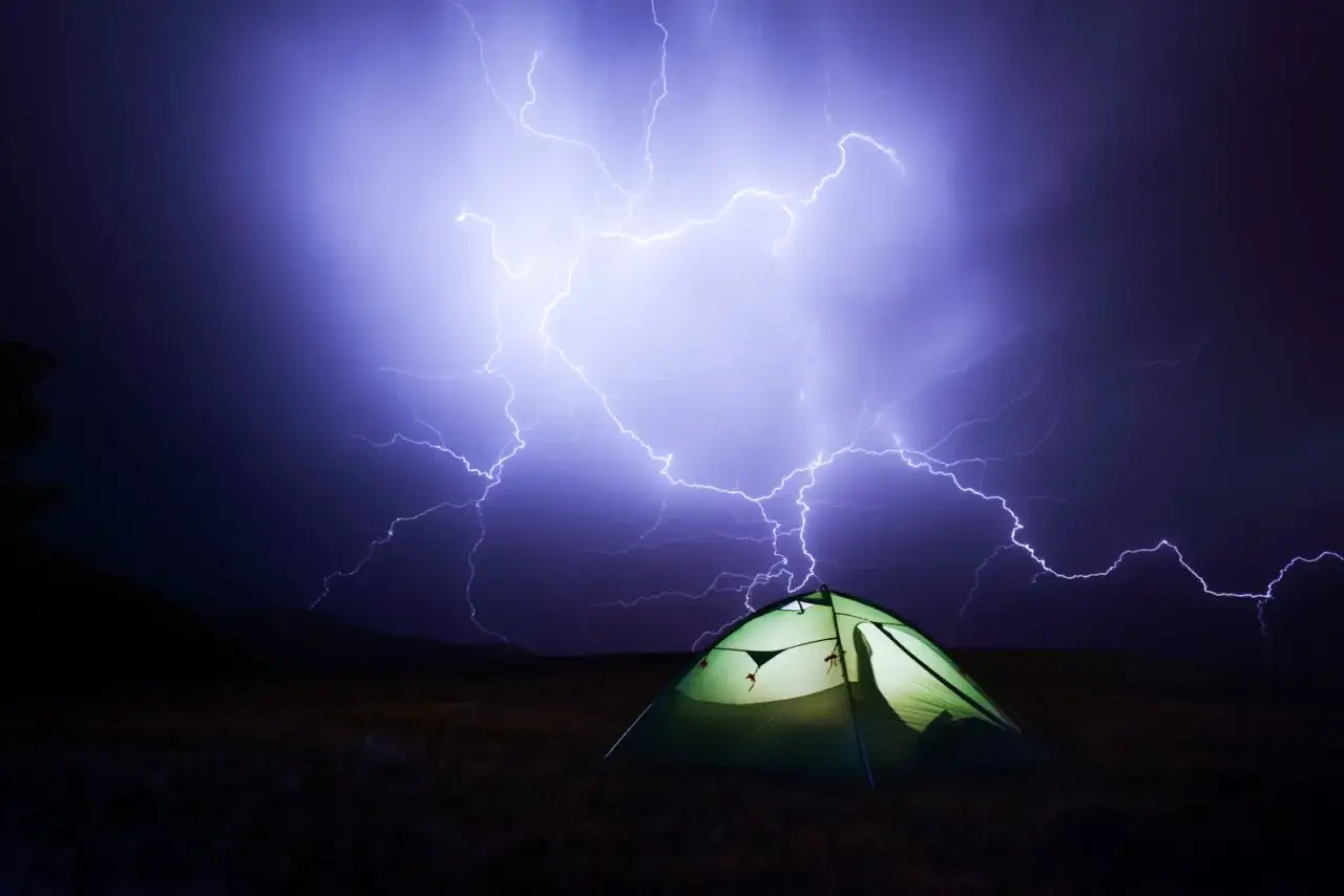 Ein grünes Zelt steht auf einer Wiese, während ein dramatisches Gewitter mit Blitzen den Himmel erhellt.
