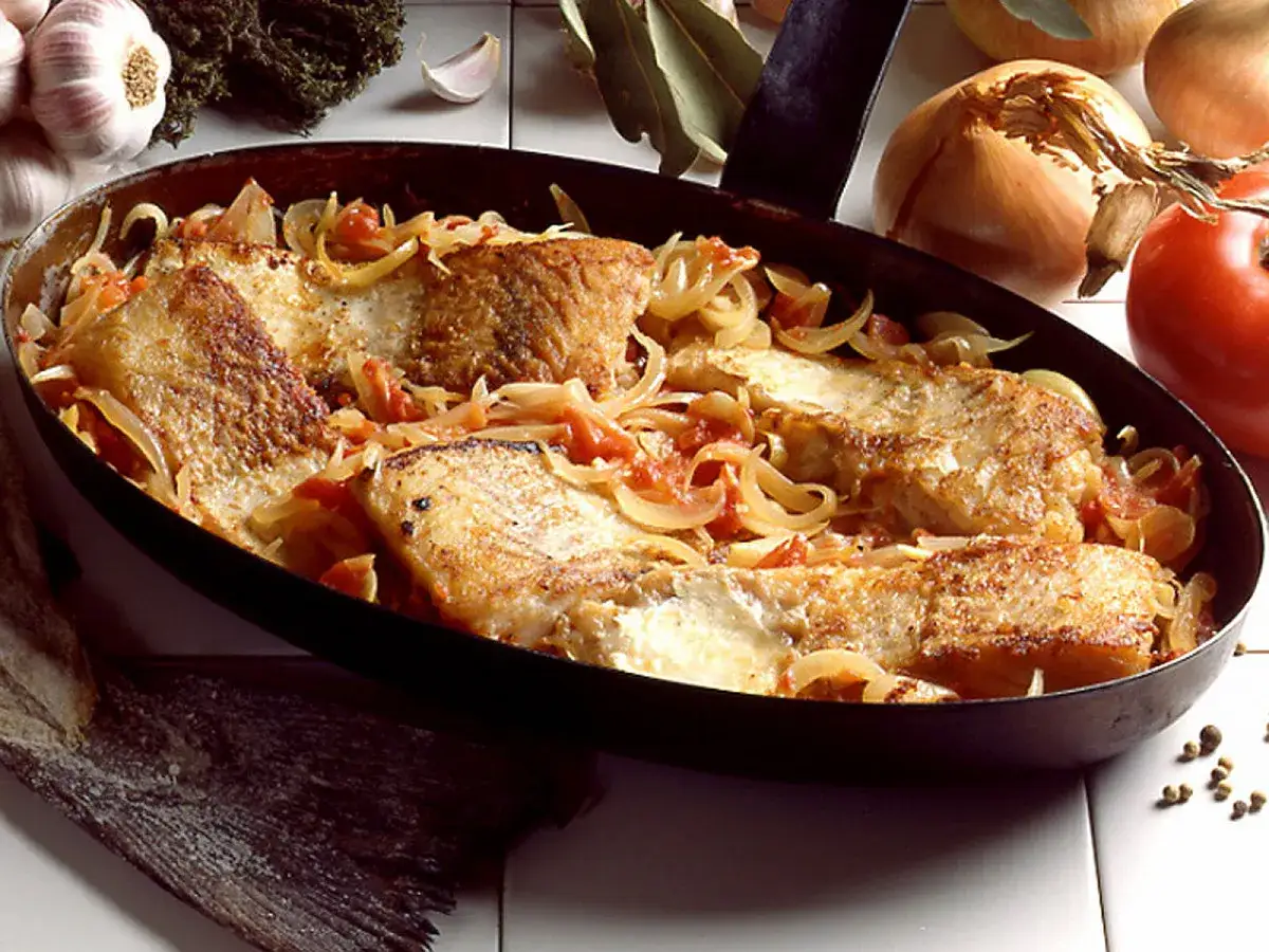 Morue à la lyonnaise, des filets de poisson dorés mijotant avec des oignons et des tomates dans une poêle.