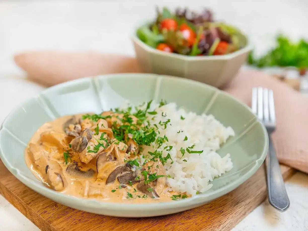 En krämig kyckling stroganoff med champinjoner och ris, toppad med persilja. En sallad syns i bakgrunden.