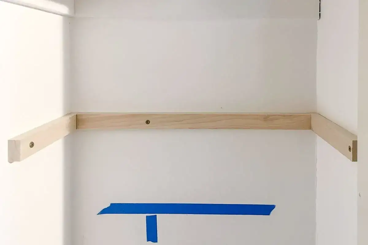 Wooden supports are being installed for DIY closet shelves. Blue tape marks a level line on the white wall.