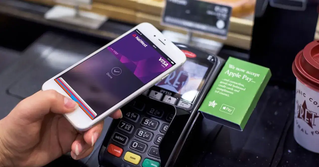 Jak płacić Apple Pay? Telefon zbliżony do terminala, na ekranie potwierdzenie transakcji. Obok zielony znak informujący o akceptacji Apple Pay.
