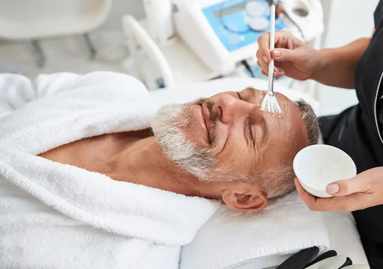 Un homme d'âge mûr, le visage rasé, reçoit un soin du visage. Une esthéticienne applique un produit sur son front avec un pinceau.