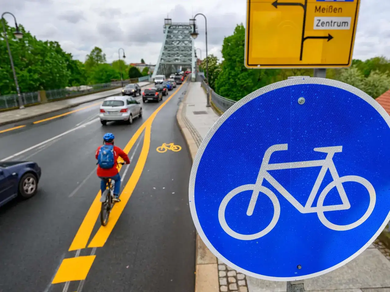 Radfahrer auf einem markierten Radweg, der über eine Brücke führt. Ein blaues Schild mit Fahrradsymbol (vz 237) und ein gelbes Ortsschild sind zu sehen.