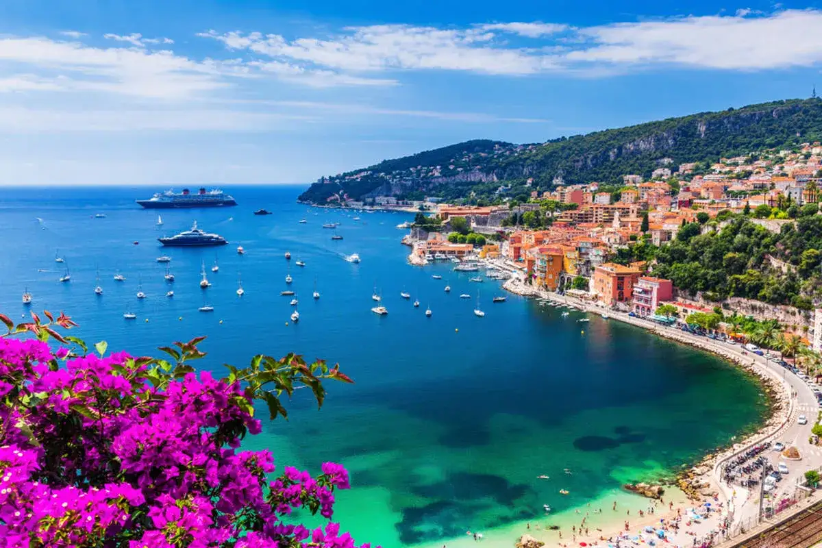 Vista de un pueblo costero en la costa atlántica francesa, con barcos en el mar y flores moradas en primer plano.