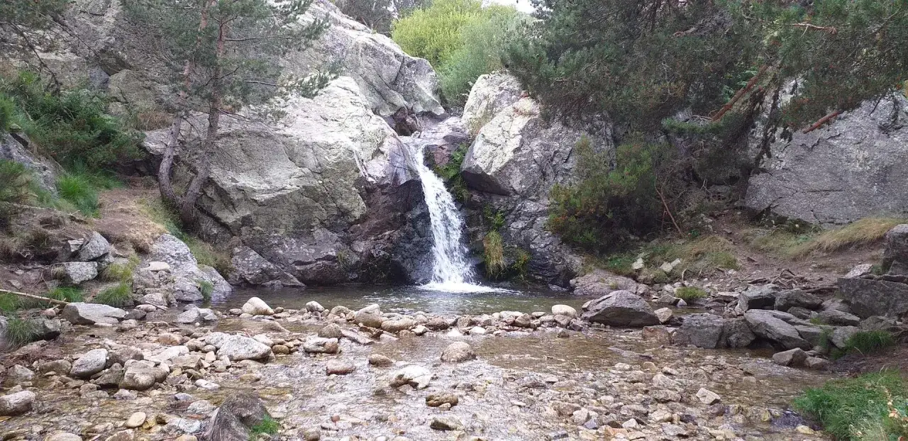 Cascada en el embalse de Navacerrada, rodeada de rocas y vegetaci&oacute;n. El agua cae sobre un lecho pedregoso.