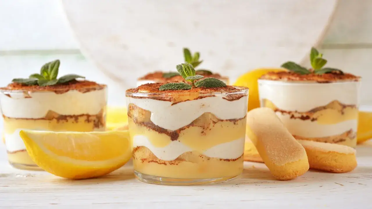 Delizioso tiramisù al limone in coppette, guarnito con menta e cacao. Accanto, spicchi di limone e savoiardi.