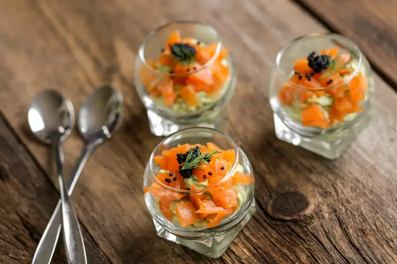 Trois verrines avocat saumon, garnies de caviar et d'aneth, sur une table en bois. Deux cuillères argentées sont posées à gauche.