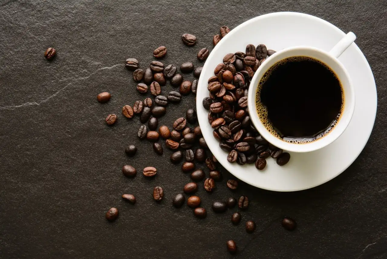 En kopp med m&ouml;rkt java kaffe st&aring;r p&aring; en vit tefat, omgiven av rostade kaffeb&ouml;nor p&aring; en m&ouml;rk yta.