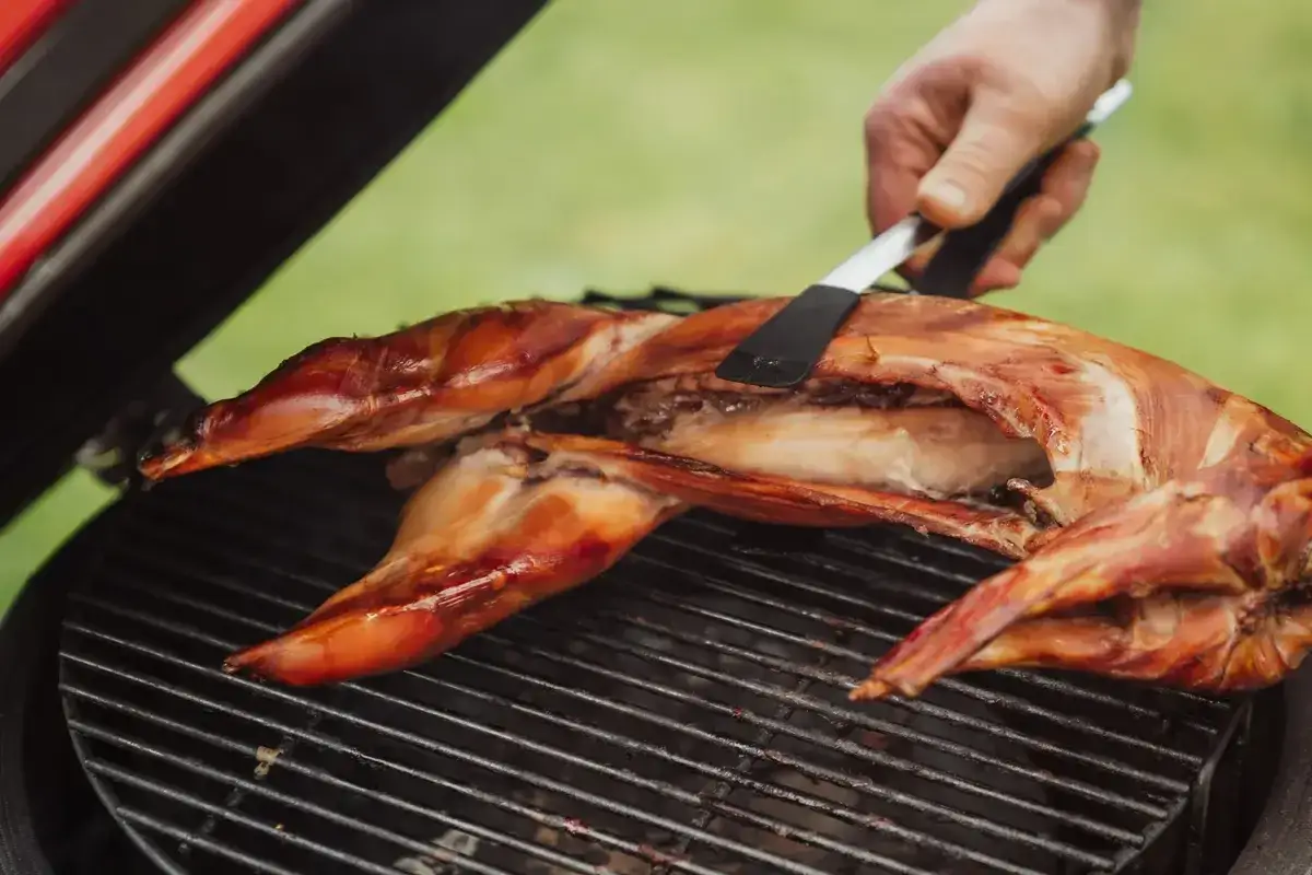 Un lapin entier est cuit sur un barbecue, sa peau dor&eacute;e et croustillante. Une main utilise une spatule pour le retourner.