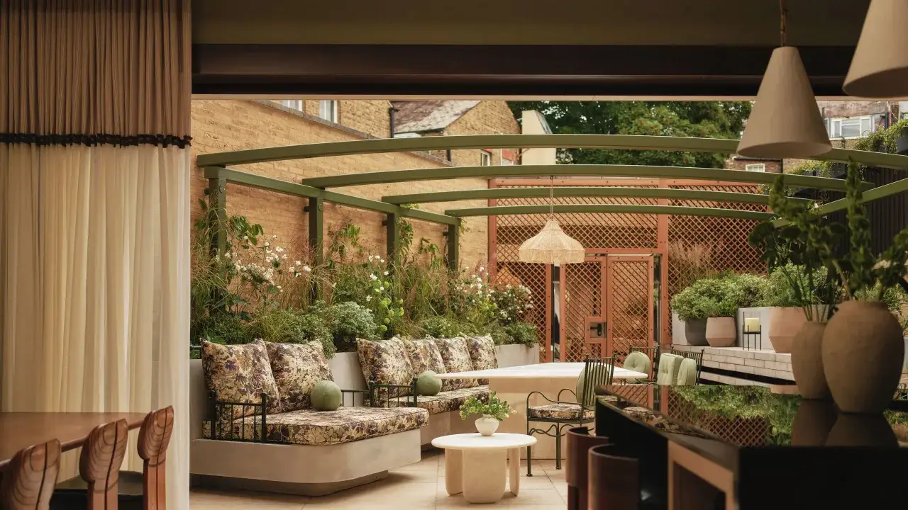 Relaks na tarasie z meblami w kwiaty i zielenią. Metalowa pergola w kolorze szałwiowym otacza przestrzeń.